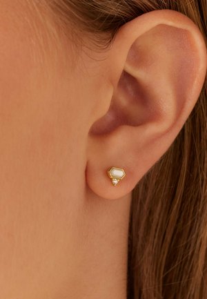 Gouden hexagonale stud oorbel met een witte inlage en een kleine heldere edelsteenaccent, gefotografeerd tegen een close-up van een oor.