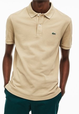 Mężczyzna ubrany w beżową koszulkę polo z krótkim rękawem z zielonym logo krokodyla, w połączeniu z ciemnozielonymi spodniami, ręce w kieszeniach.