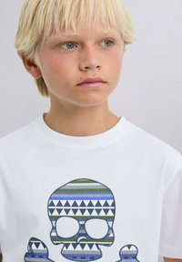 Valkoinen puuvillainen t-paita, jossa on kuvioitu sininen ja vihreä pääkallo graafinen design geometrisine muotoineen. Klassinen pyöreä kaula-aukko ja lyhyet hihat.