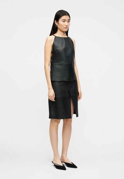 Γυναίκα που στέκεται, φορώντας ένα αμάνικο μαύρο δερμάτινο τοπ και αντίστοιχη φούστα μήκους έως το γόνατο με σχίσιμο μπροστά, συνδυασμένα με μαύρα παπούτσια slingback με μυτερή μύτη.