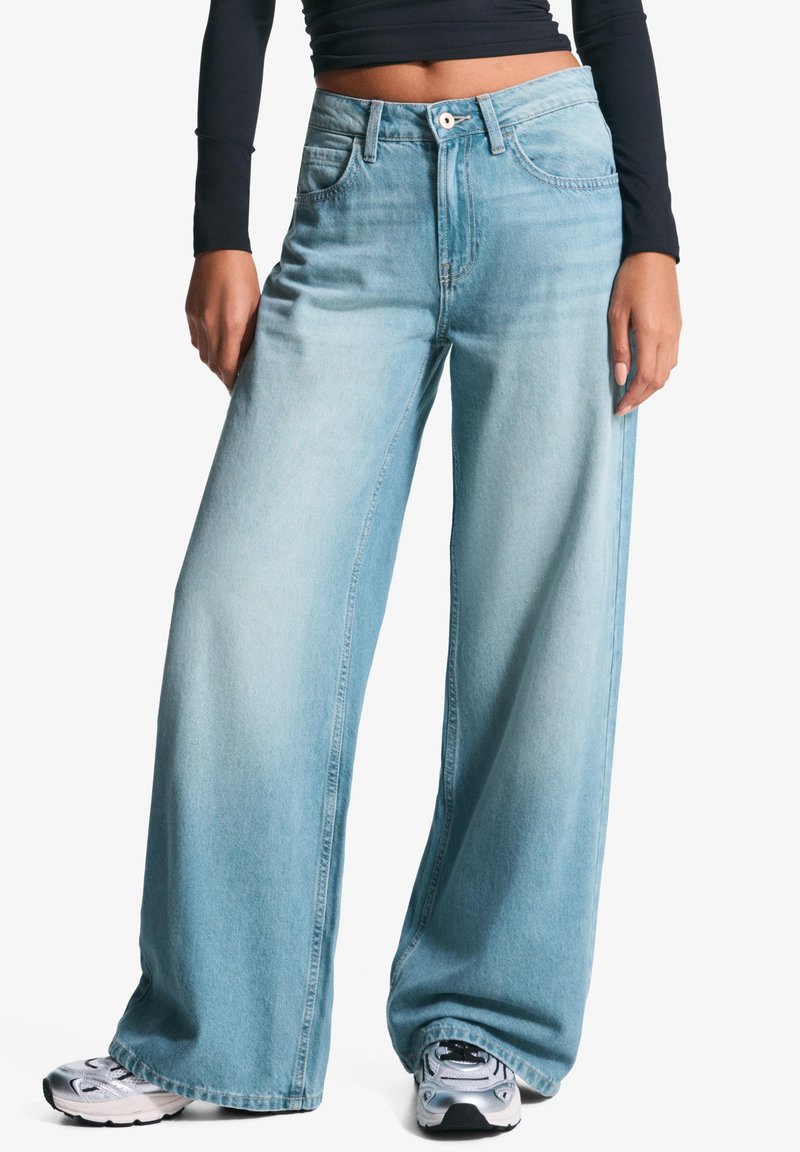Široké světlé modré denimové džíny s mírným praním, vysokým pasem, velkými kapsami a jemným vyblednutím. V kombinaci se stříbrnými teniskami.