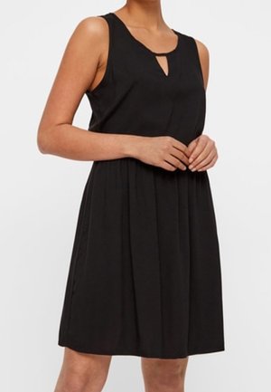 Czarna, bezrękawnikowa sukienka do kolan z małym dekoltem w kształcie litery V, noszona przez osobę ze złożonymi rękami z przodu.