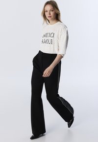 Top bianco a maniche lunghe con texture e la scritta "MERCI AMOUR" in nero, abbinato a pantaloni neri a vita alta con strisce bianche sui lati.