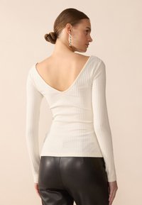 Long-sleeve, ribbed top in off-white with a deep V-back design. Paired with black, form-fitting pants for a sleek contrast.