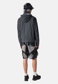 Dunkelgraue Windjacke mit Kapuze, kombiniert mit shorts in Schwarz und Grau mit geometrischem Muster und schwarzen Turnschuhen. Ein Bucket Hat rundet das Outfit ab.