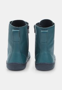 Un paio di stivaletti in pelle color teal con suole nere e cerniere sul lato interno, visti da dietro su uno sfondo bianco.