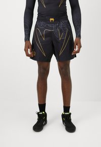Mann in schwarzen Sportshorts mit goldener Schlangendesign, langen schwarzen Kompressionsärmeln und schwarzen Turnschuhen mit neongrünen Akzenten.