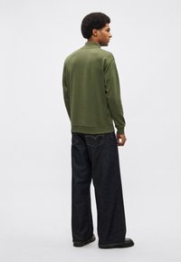 Oliiviroheline kaelusega pullover, mida kantakse koos laia säärega tume teksapükstega. Kangas tundub sile ja pehme. Mustad kingad täiendavad rõivakomplekti.