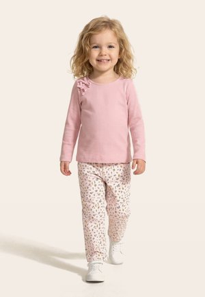 Dijete koje se smije s kovrčavom plavom kosom hoda prema naprijed, nosi ružičastu majicu dugih rukava s detaljem mašne, cvjetne hlače i bijelu obuću.