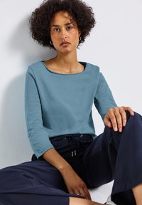 Camicia azzurra chiara a maniche lunghe con scollatura ampia, abbinata a pantaloni blu scuro. Tessuto morbido, vestibilità casual e design minimalista.