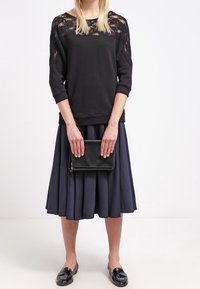 Maglione nero con dettagli in pizzo, abbinato a una gonna a pieghe blu navy. La modella tiene una piccola pochette nera. Mocassini neri lucidi completano l'outfit.
