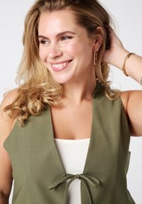 Olijfgroene mouwloze top met een striksluiting aan de voorkant, gecombineerd met een witte onderkleding. Glad materiaal en eenvoudig ontwerp.