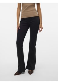 Pantalon noir évasé en tissu doux et extensible ; doté d'une taille ajustée et de détails subtils sur les coutures. Porté avec des chaussures marron à bout pointu.