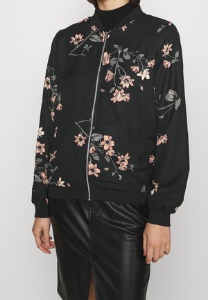 Zwarte jas met rits en roze bloemenprint, gedragen over een zwarte coltrui, gecombineerd met een zwarte leren rok met een voorsplit.