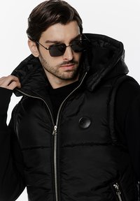 Czarna kurtka puchowa bez rękawów z zamkiem z przodu i kapturem, z błyszczącym wykończeniem, pikowanym teksturem oraz okrągłym logo na klatce piersiowej.