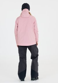 Rosa wasserdichte Jacke mit hohem Kragen und verstellbarem Kapuze, kombiniert mit dunkelgrauen Hosen im weiten Schnitt und mit strukturiertem Stoff.