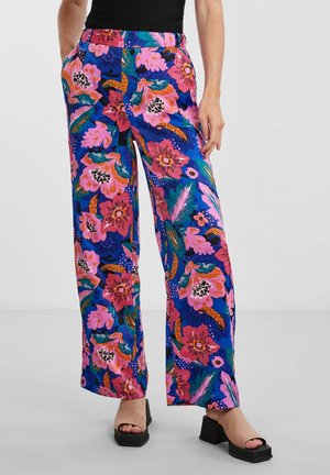 Une personne porte un pantalon à fleurs aux motifs vifs en rose, bleu et vert, associé à des sandales à plateforme noires, se tenant devant un arrière-plan neutre.