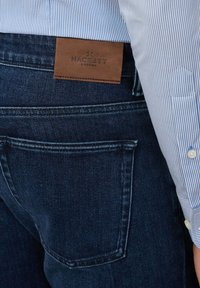 Kratak pogled na stražnji džep i kožnu oznaku branda na tamnoplavim trapericama, uz vidljiv rukav majice u plavoj i bijeloj prugici.