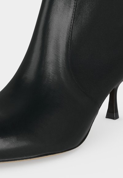 Gros plan d’une botte cheville en cuir noir avec un bout pointu et un talon haut fin et incliné sur fond blanc.