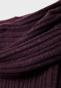 Neulos kankaalla syvän liilan sävyisenä, jossa on pystysuora ribbitekstuuri. Kangas näyttää pehmeältä ja siinä on roikkuvia taitoksia, jotka korostavat sen joustavuutta ja lämpöä.