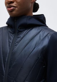 Prim-plan cu o persoană care poartă o jachetă matlasată albastru închis, cu fermoar frontal și glugă, arătând detaliile pieptului superior și gâtului.