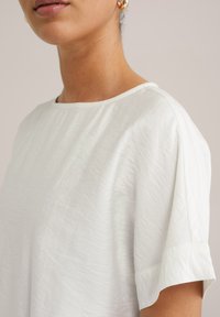Bílý volný top s měkkou texturovanou tkaninou. Má kulatý výstřih a krátké vyhrnuté rukávy. Jednoduchý a minimalistický design.