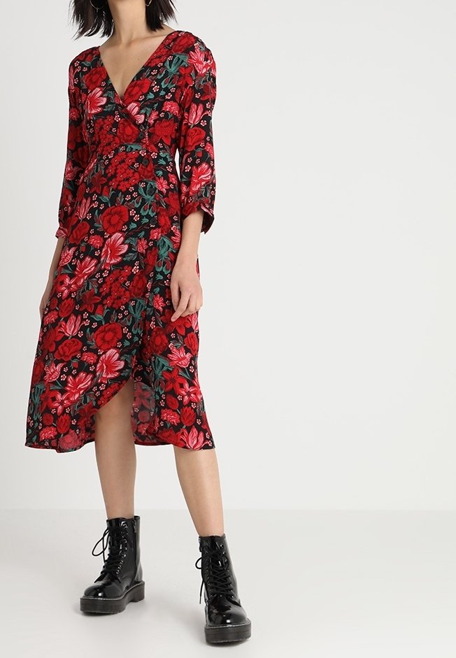 Femme portant une robe portefeuille fleurie rouge et noire avec des manches trois-quarts et des bottines noires à lacets, debout sur un fond uni.