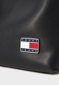 Fekete textúrájú anyag, ezüst színű "TOMMY JEANS" logó táblával, piros és fehér díszítéssel négyzetes háttéren.