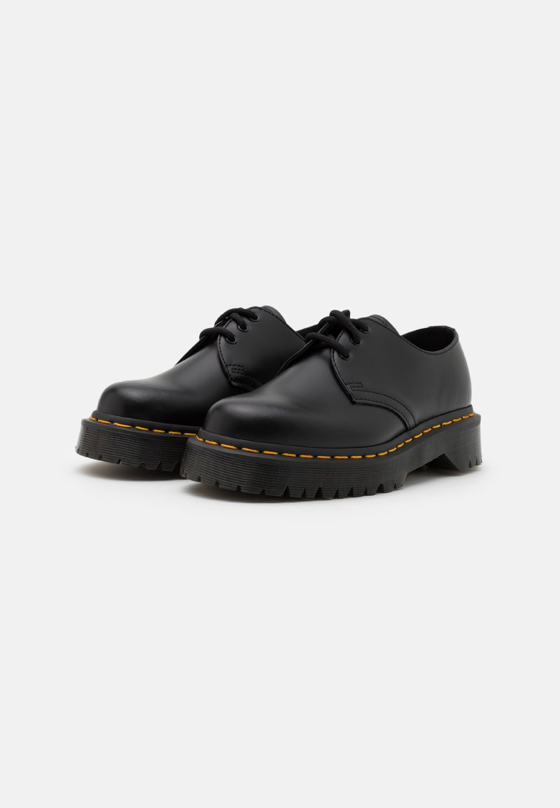 Scarpe in pelle nera con punta rotonda, design con lacci, suola in gomma testurizzata e cucitura gialla a contrasto lungo il bordo.