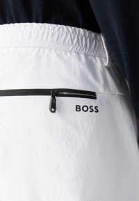 Λευκά σορτς με ελαστική μέση, μαύρη λεπτομέρεια με φερμουάρ στην τσέπη και μαύρο λογότυπο "BOSS" στο πλάι. Υφασμάτινη υφή.