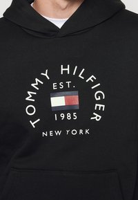 Sudadera negra con capucha que presenta un logo circular con "TOMMY HILFIGER", est. 1985, y un pequeño diseño de bandera en rojo, blanco y azul. Tejido suave, bolsillo tipo canguro.