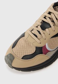 Beige sneaker med mesh-paneler, svarta accenter och texturerad gummisula. Snörningen är ljusbeige med en kontrasterande vinröd detalj.