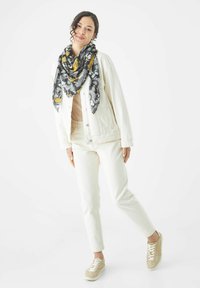 Witte denim jas over een beige top, gecombineerd met witte broeken en lichtbeige sneakers. Een zwarte sjaal met een geel en grijs patroon voegt detail toe.