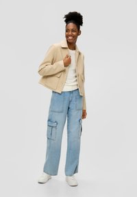 Beige Cropped-Jacke mit Kragen und Fronttaschen, kombiniert mit hellblauen, weit geschnittenen Cargo-Hosen mit großen Seitentaschen, getragen mit weißen Sneakers.