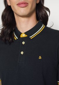 Pólo preto feito de tecido texturizado, com um colarinho amarelo com riscas brancas, botões amarelos e um pequeno logótipo bordado.