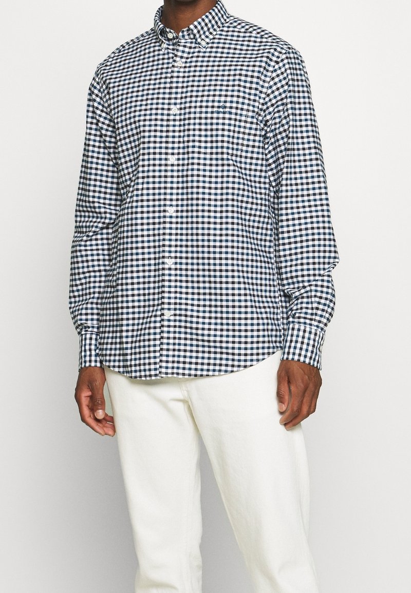Homme portant une chemise à manches longues à carreaux noirs et blancs avec un pantalon blanc sur un fond blanc uni.