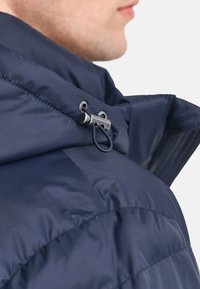 Marineblå dunjakke med høj krave og metal-lukning. Har quiltet tekstur og glat, skinnende stoffinish.