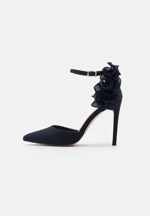 Zapato negro de tacón alto con punta en pico, tira en el tobillo y detalle decorativo de tela fruncida en la parte trasera.