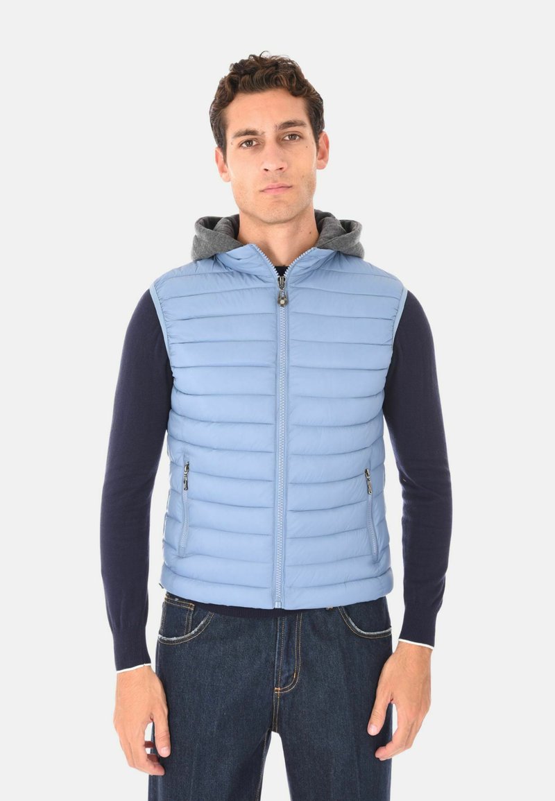Uomo che indossa un gilet smanicato trapuntato blu chiaro con cappuccio grigio sopra una maglietta a maniche lunghe blu navy e jeans scuri, in piedi su uno sfondo semplice.