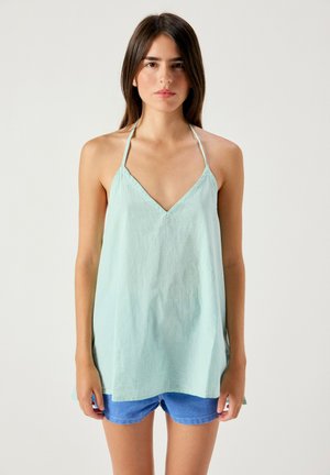 Młoda kobieta z długimi brązowymi włosami, ubrana w jasn Ziee halter top i niebieskie szorty, stojąca na jednolitym białym tle.