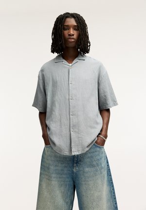 Camicia a maniche corte in lino grigio chiaro con colletto, caratterizzata da una chiusura frontale con bottoni e una vestibilità morbida, abbinata a pantaloni in denim blu sbiaditi e larghi.