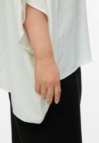 Biały, lekki top o fakturowanej powierzchni i luźnych rękawach, zestawiony z czarnymi spodniami. Widoczna ręka spoczywająca po boku.