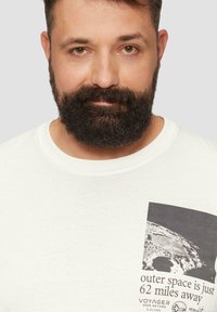 Witte katoenen t-shirt met een zwart-wit afbeelding van de Maan en de tekst "de ruimte is slechts 100 kilometer verderop" aan de linkerkant.