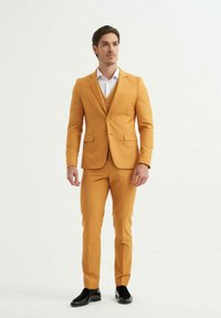 Costume jaune avec une coupe ajustée, veste à un seul bouton et pantalon assorti. Chemise blanche en dessous, chaussures noires complètent la tenue.