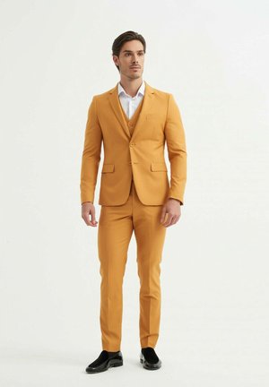 Costume jaune avec une coupe ajustée, veste à un seul bouton et pantalon assorti. Chemise blanche en dessous, chaussures noires complètent la tenue.