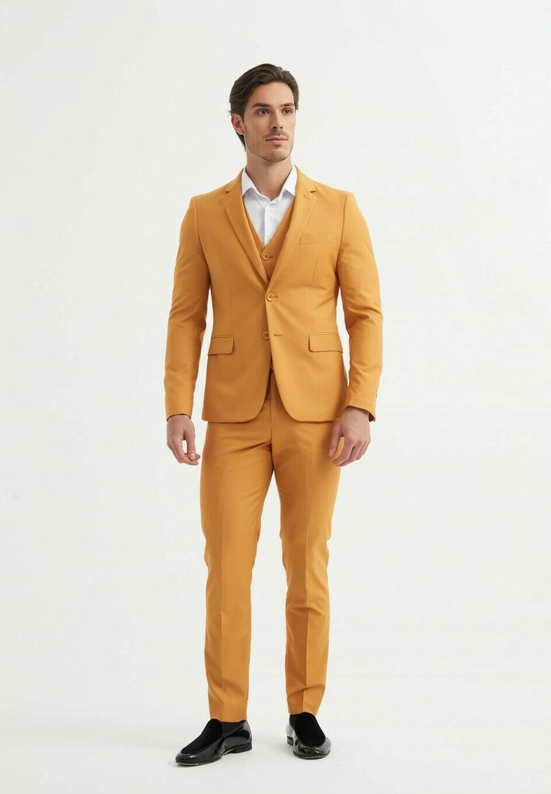 Costume jaune avec une coupe ajustée, veste à un seul bouton et pantalon assorti. Chemise blanche en dessous, chaussures noires complètent la tenue.