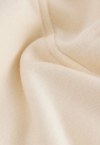 Tissu de couleur crème à surface douce et texturée, présentant des variations subtiles et des plis, indiquant un matériau tricoté léger.