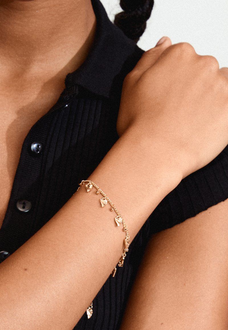 Gouden charm armband met hartvormige hangers, voorzien van een delicate ketting en getextureerde accenten. Gedragen op een gebruinde arm tegen een zwarte top.