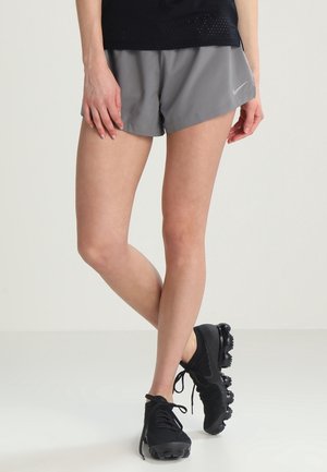 Osoba nosząca szare spodenki Nike, czarne buty sportowe i czarny top, stojąca ze skrzyżowanymi nogami na jednolitym tle.