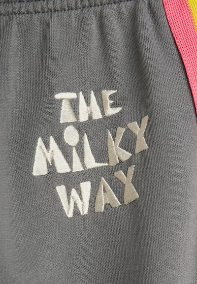 Γκρι ύφασμα με κεντημένο λευκό κείμενο "THE MILKY WAY" και ροζ και κίτρινη ρίγα λεπτομέρειας στη δεξιά πλευρά.